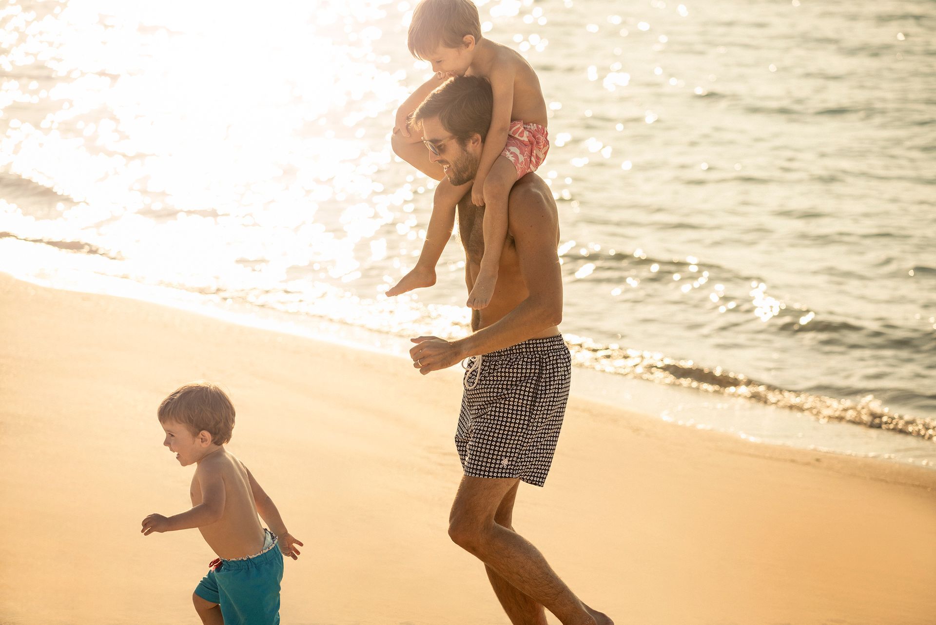Father carrying a child on his shoulders while another child runs on a beach.