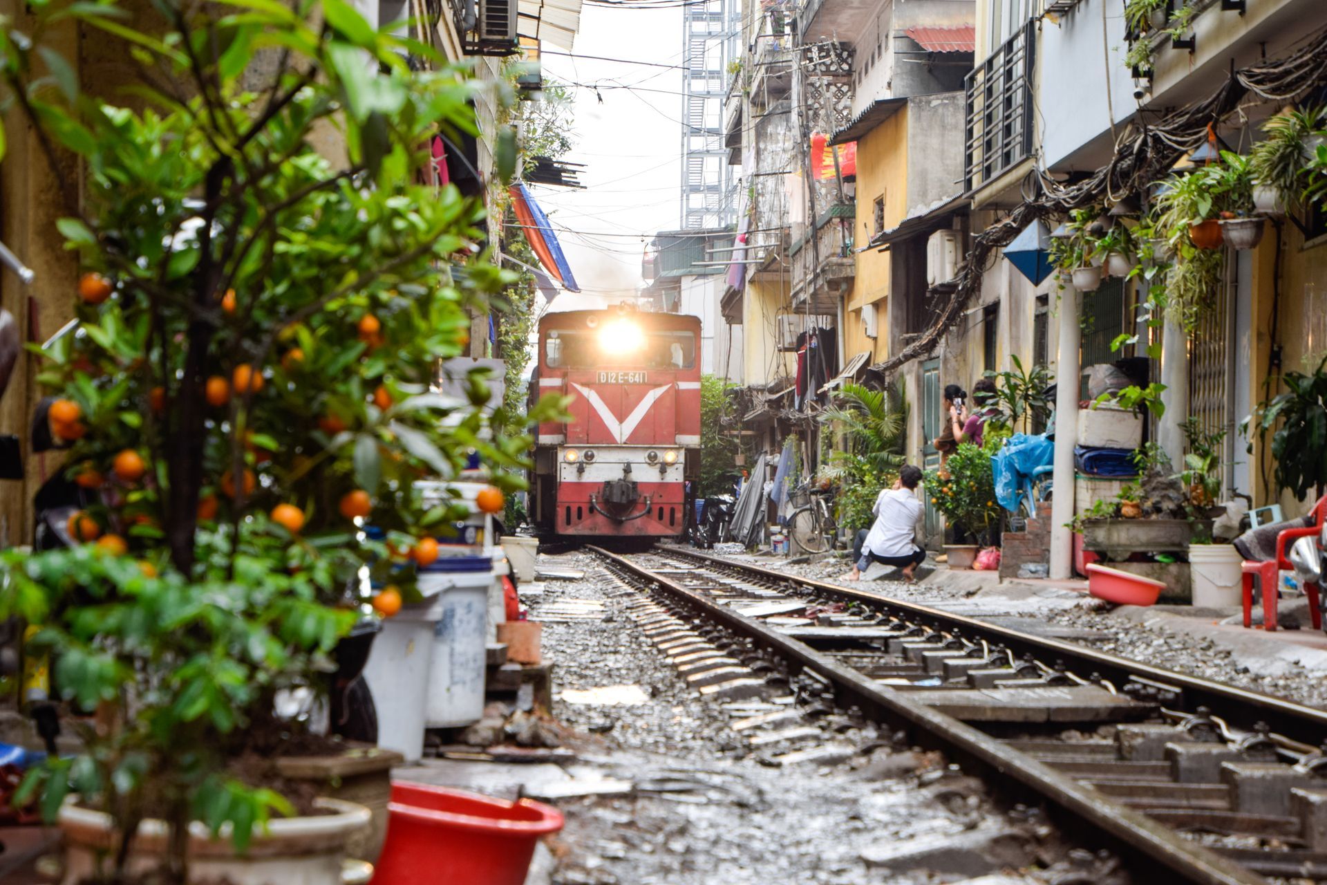 Train approaching narrow alleyway with buildings, trees, and people.