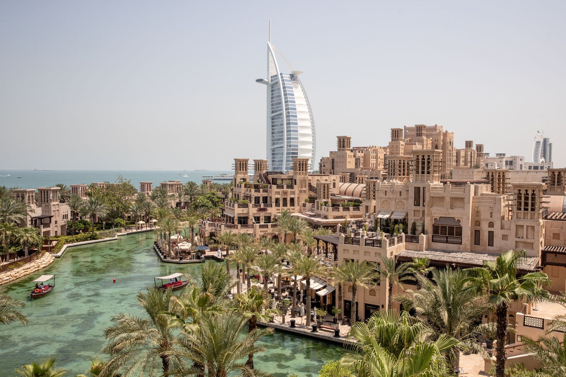 Dubai cityscape with Burj Al Arab hotel and waterway, featuring beige buildings, palm trees, and boats.