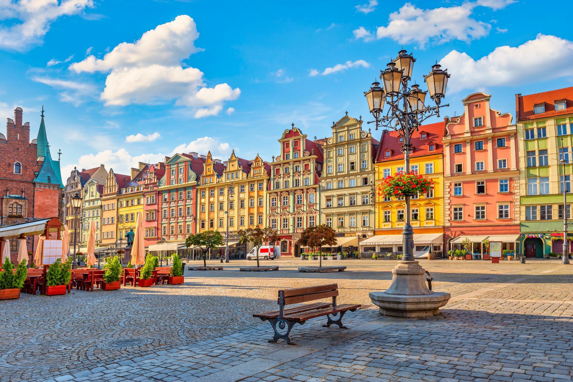 Colorful buildings surround a cobblestone square in a sunny city. A bench and lamppost are in the foreground.
