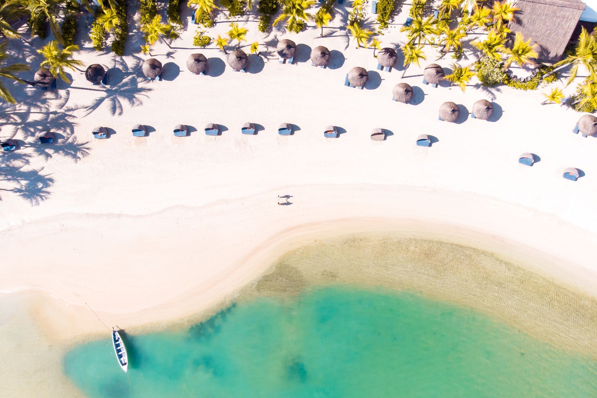 Aerial view of a white sand beach with turquoise water. Palm trees and thatched umbrellas line the shore.