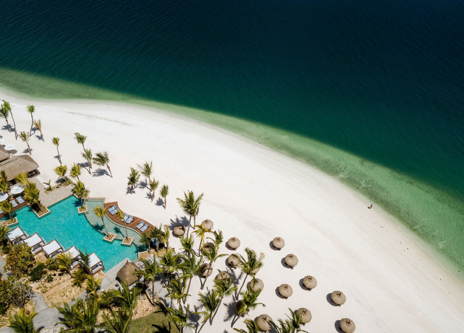 Aerial view of a white sand beach with a turquoise pool and dark blue ocean. Palm trees and beach umbrellas.