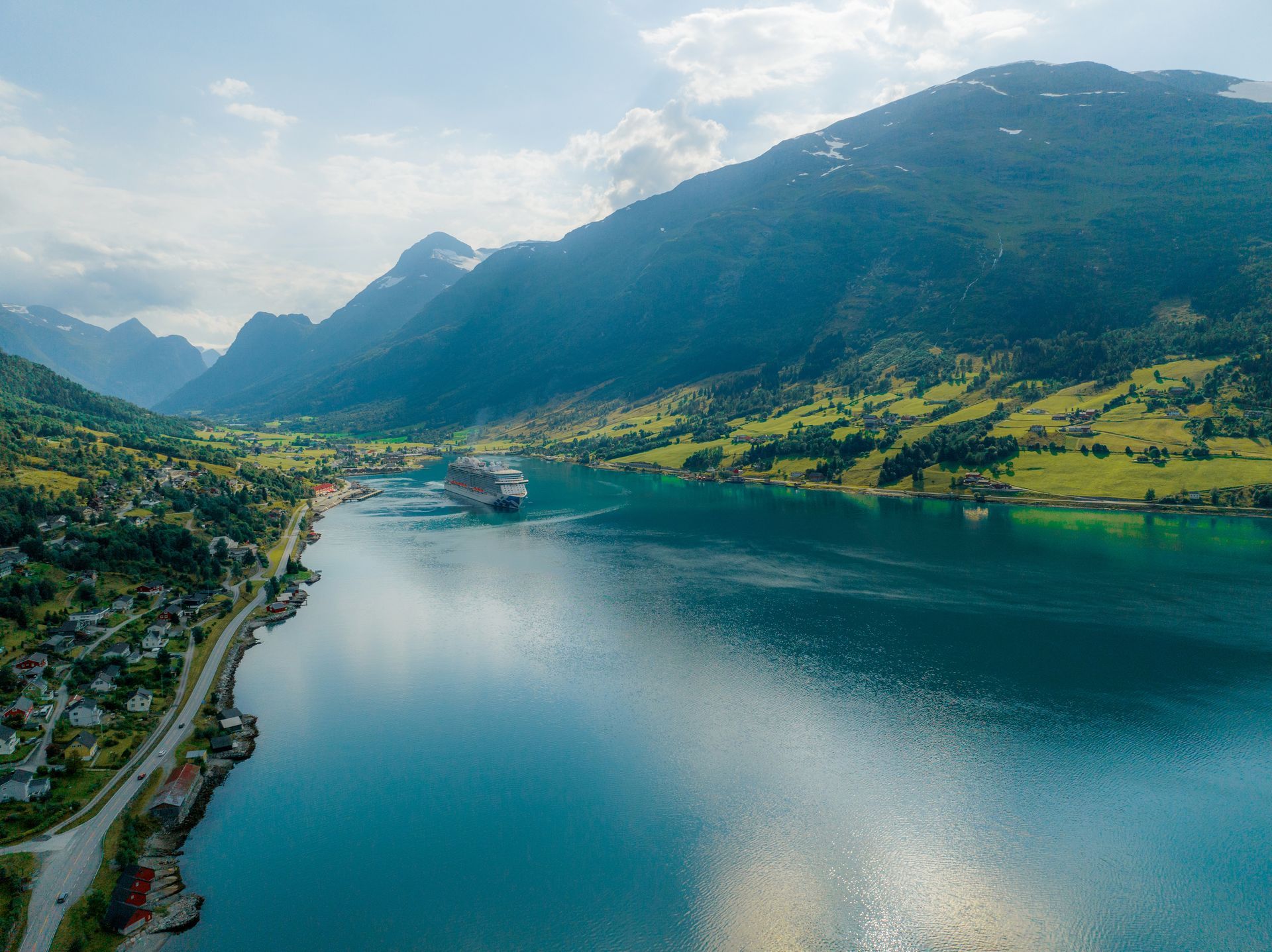Cruise ship docked in a turquoise fjord with green hills and snow-capped mountains in the background.