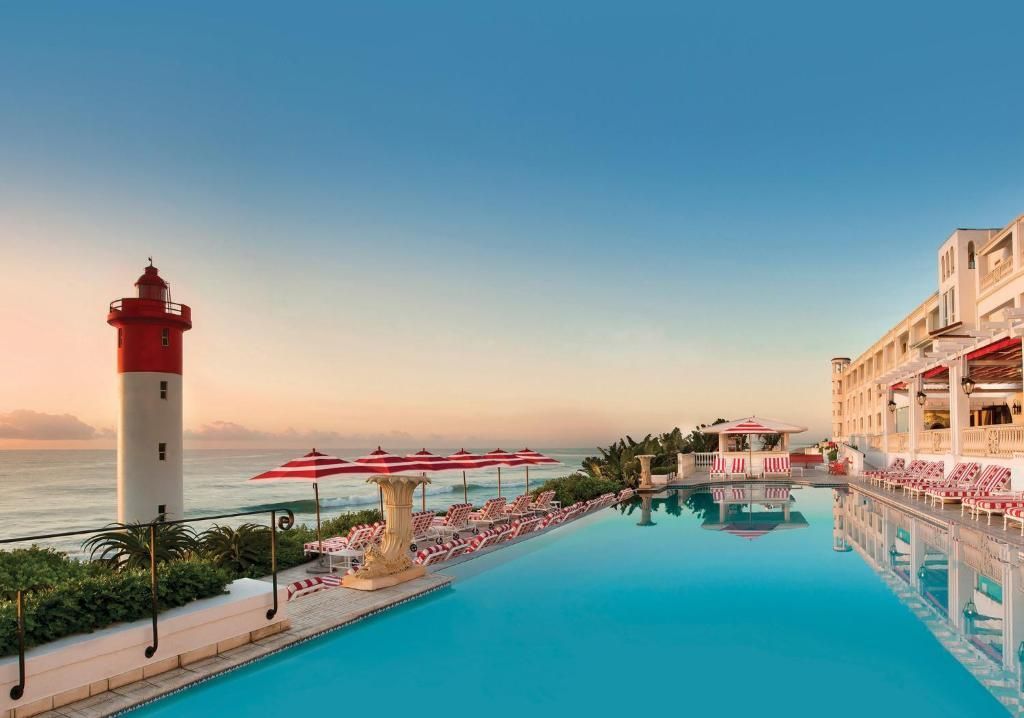 Infinity pool at a coastal resort, featuring a lighthouse, and building with balconies under a blue sky.