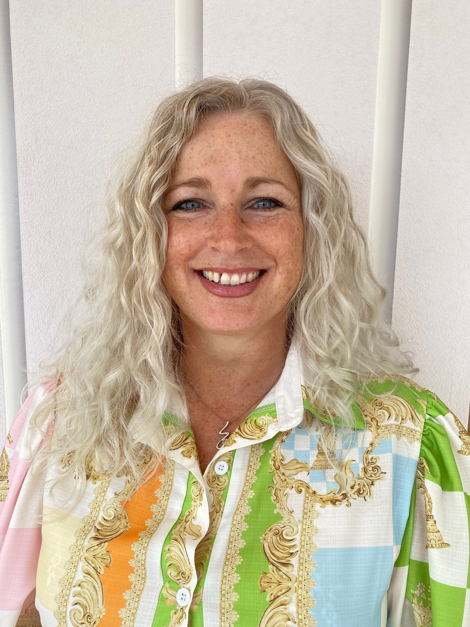 Woman with curly gray hair, smiling, wearing a colorful patterned shirt, in front of a white wall.