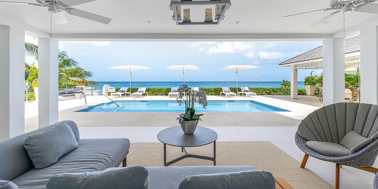 Outdoor patio with a pool and ocean view. Grey furniture, white pillars, and blue sky.