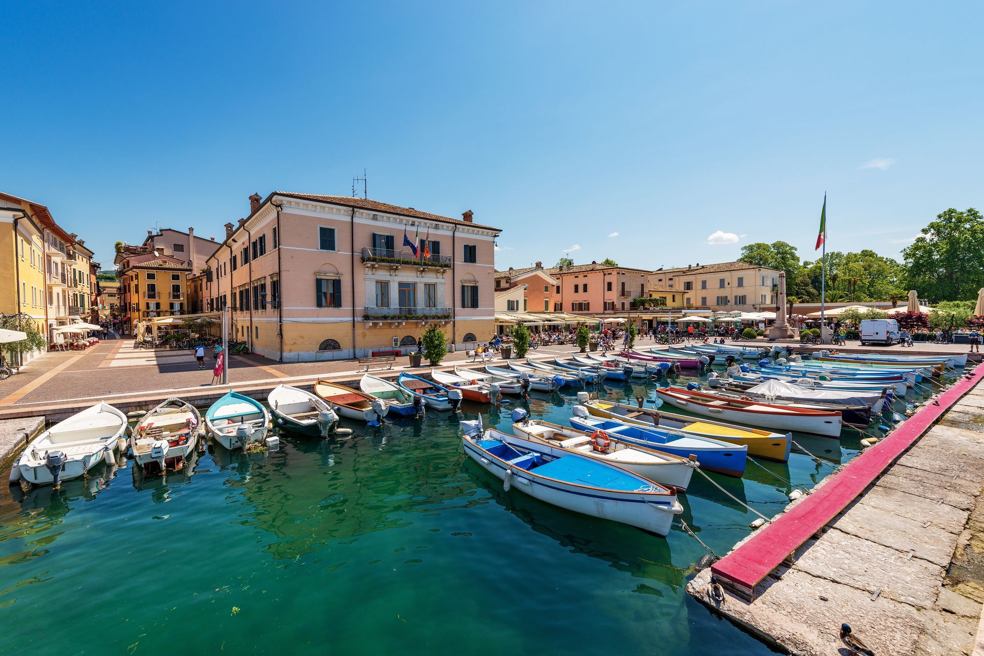 Boats docked in a harbor, pastel buildings, sunny day, Italian flag, waterfront town.