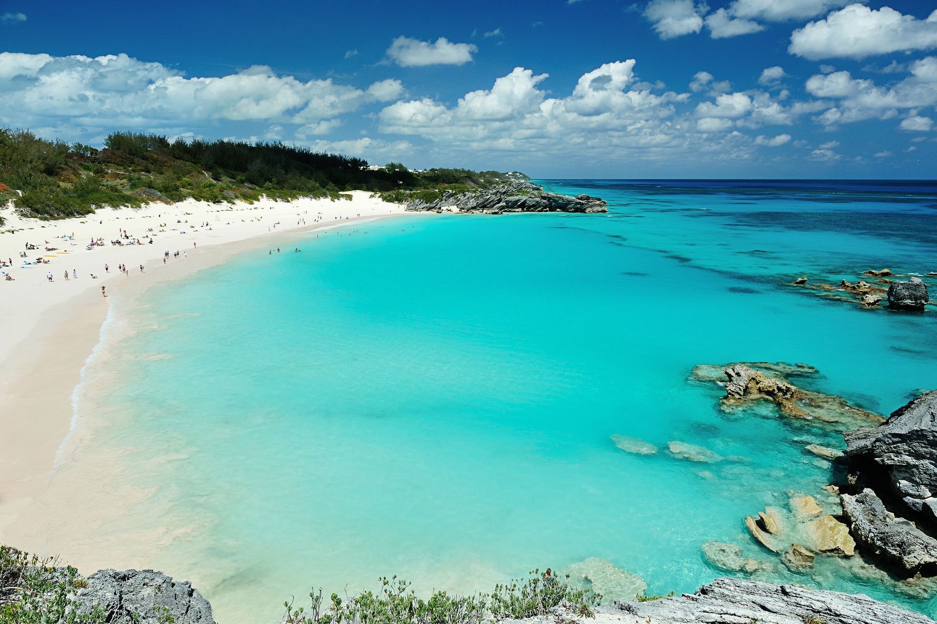 Turquoise water at a white sand beach with people, trees, and blue sky.