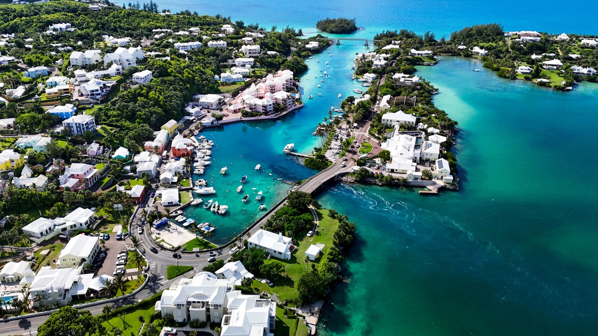 Aerial view of a coastal town with buildings, boats, and a bridge over turquoise water. Lush green trees surround the area.