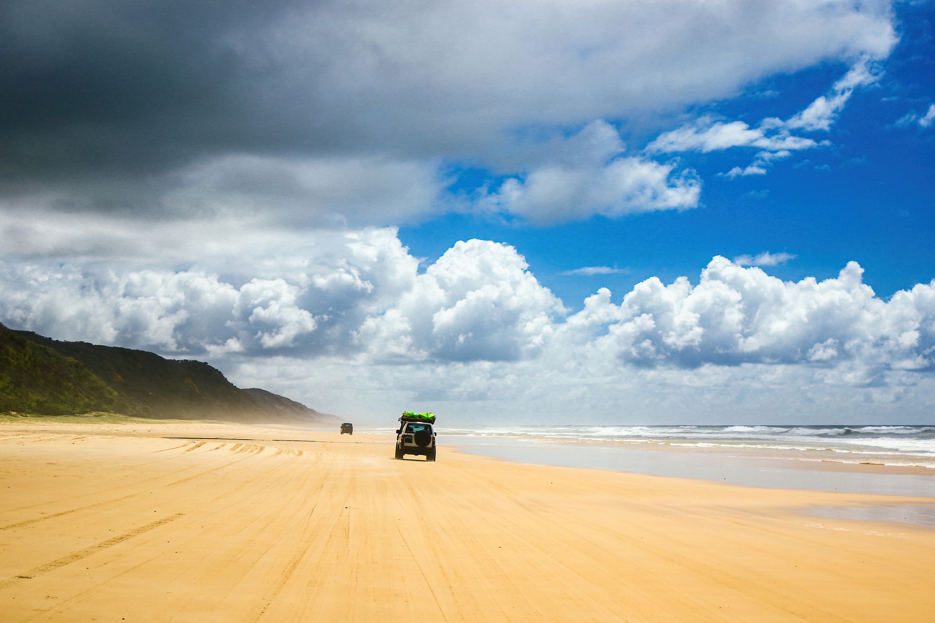 Beach with a car driving on the sand under a bright blue sky with puffy white clouds.