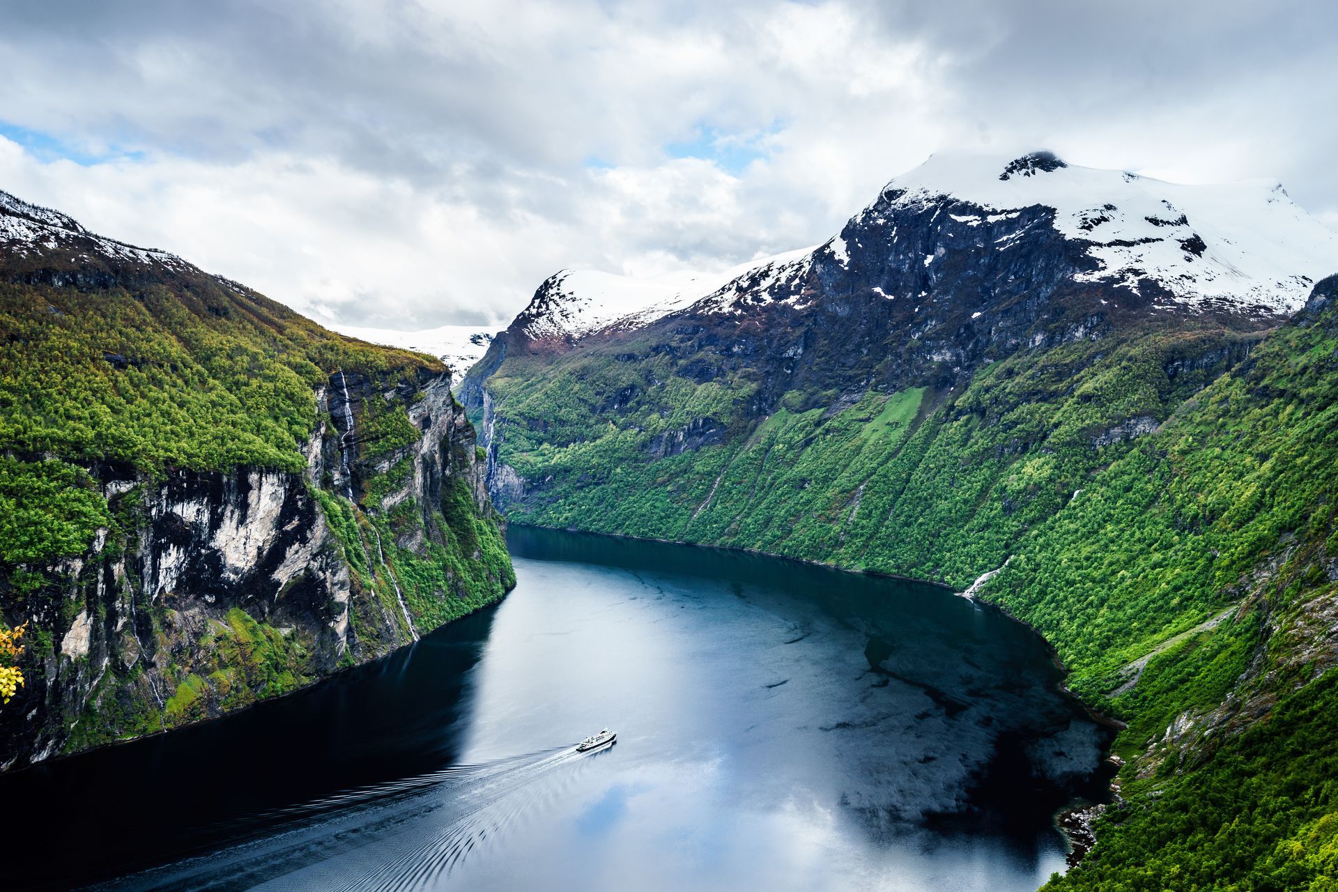 Fjord with boat, flanked by green cliffs, snow-capped mountains under cloudy sky.