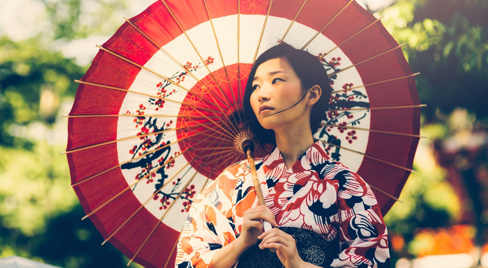 Woman in kimono holds a red and white parasol, outside, looking off-camera.