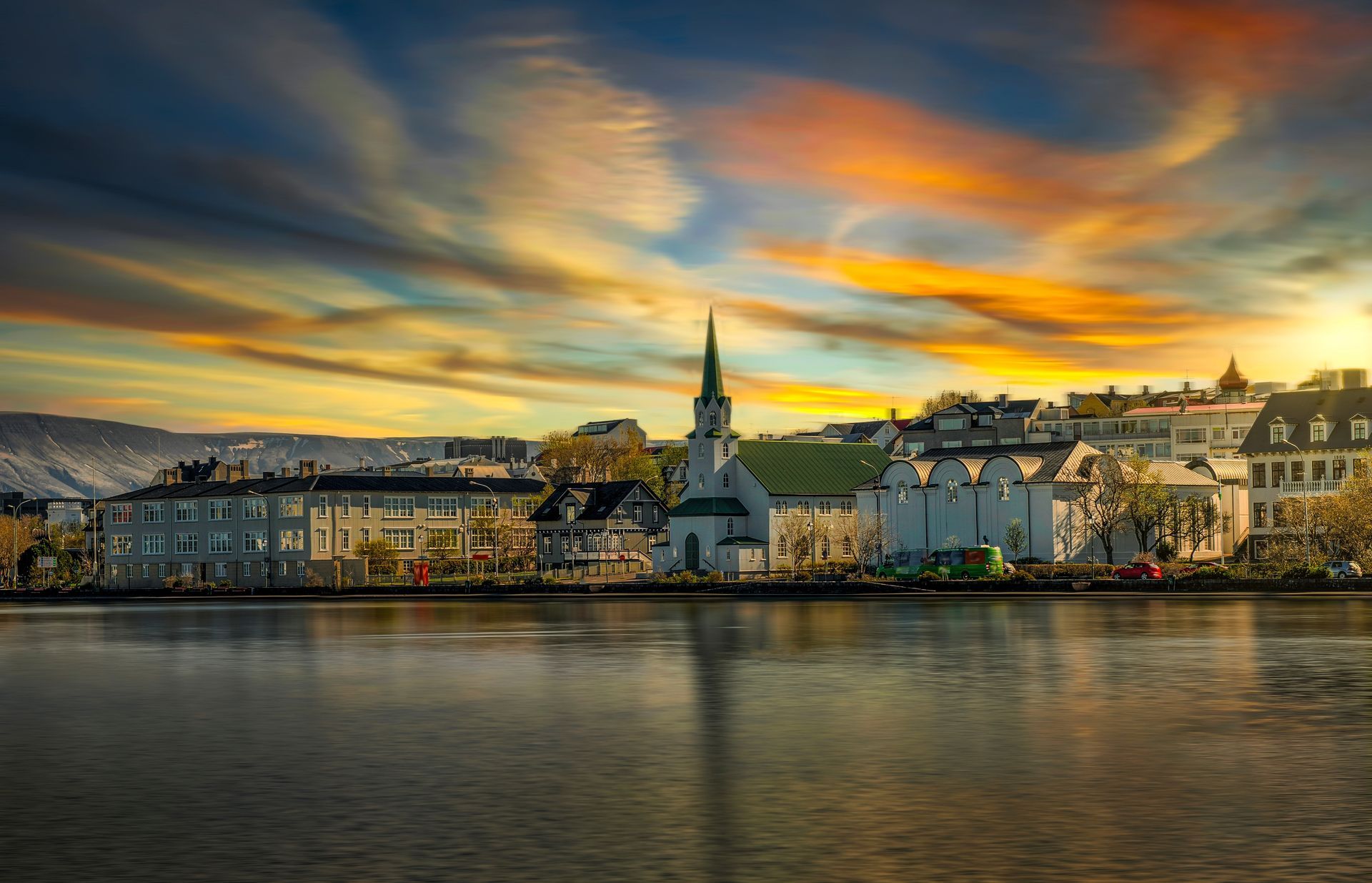 City skyline at sunset reflected in water, church steeple prominent, vibrant sky of orange and blue.