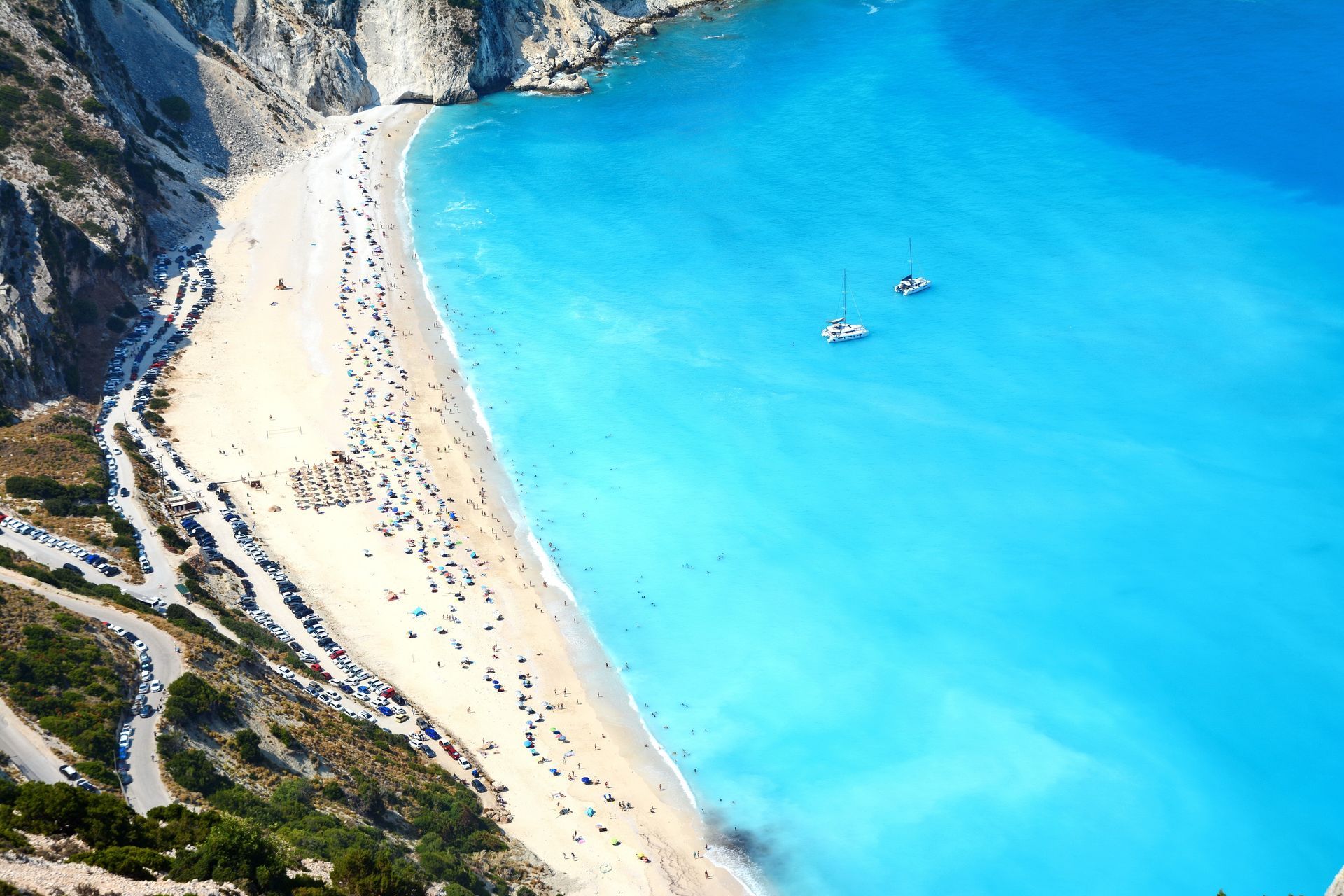 Beach with turquoise water, white sand, and people. Two boats float in the sea.