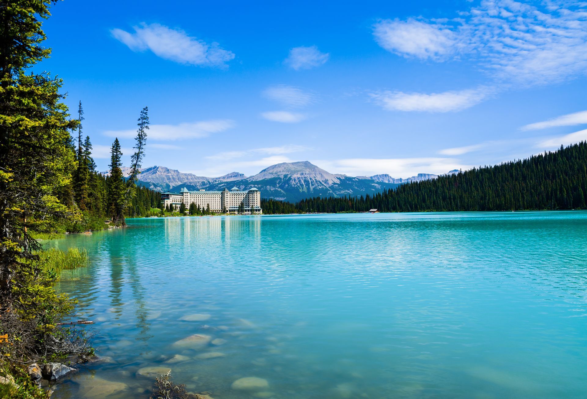Turquoise lake with mountains and a large building under a bright blue sky.