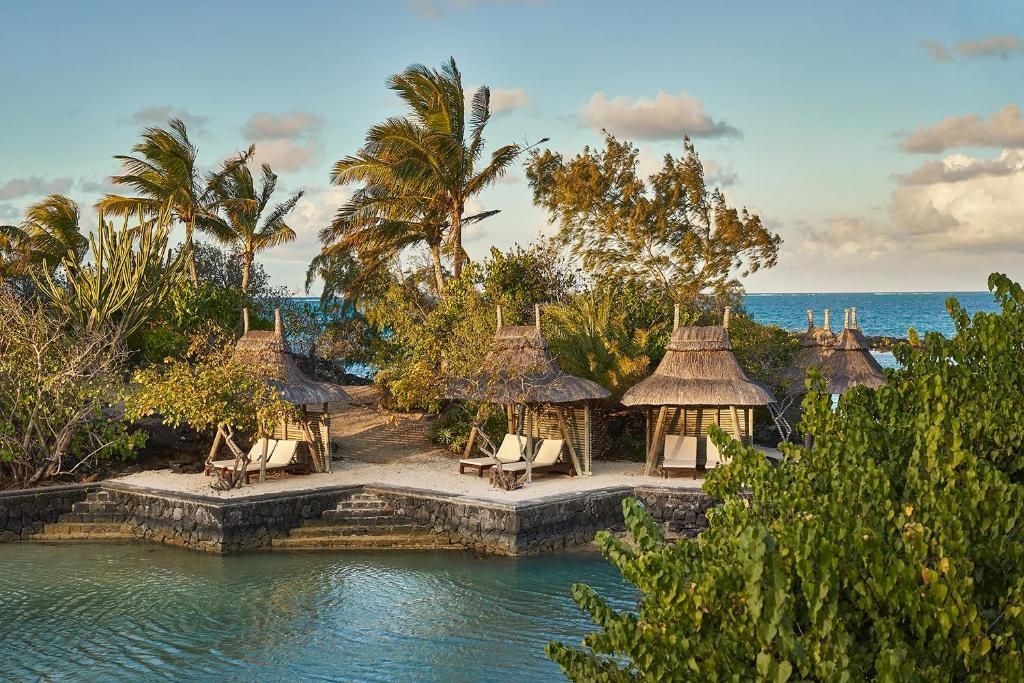 Tropical scene: thatched roof cabanas with lounge chairs on a small island, near turquoise water, under a blue sky.