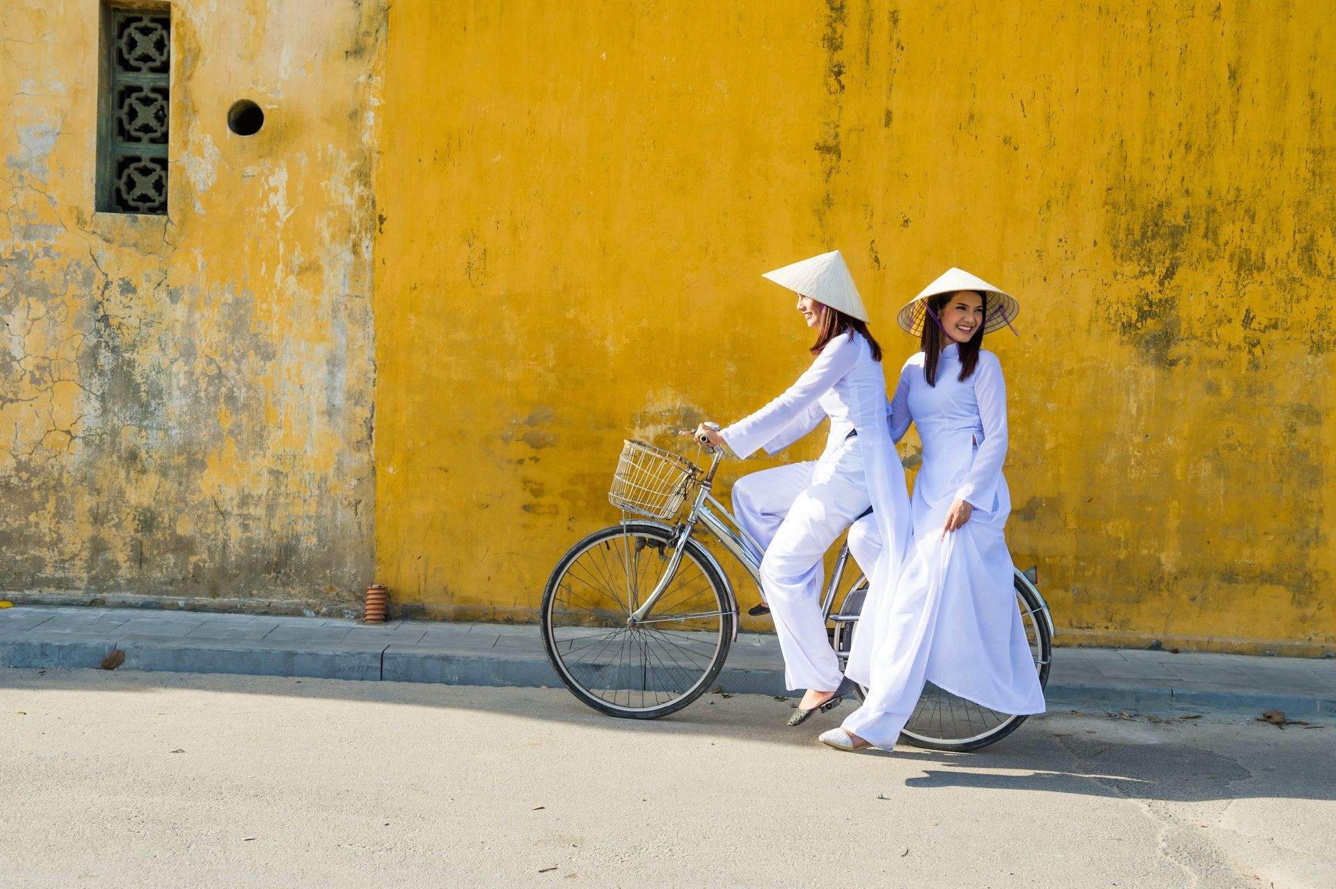 Two women in white áo dài and conical hats next to a bicycle against a yellow wall.