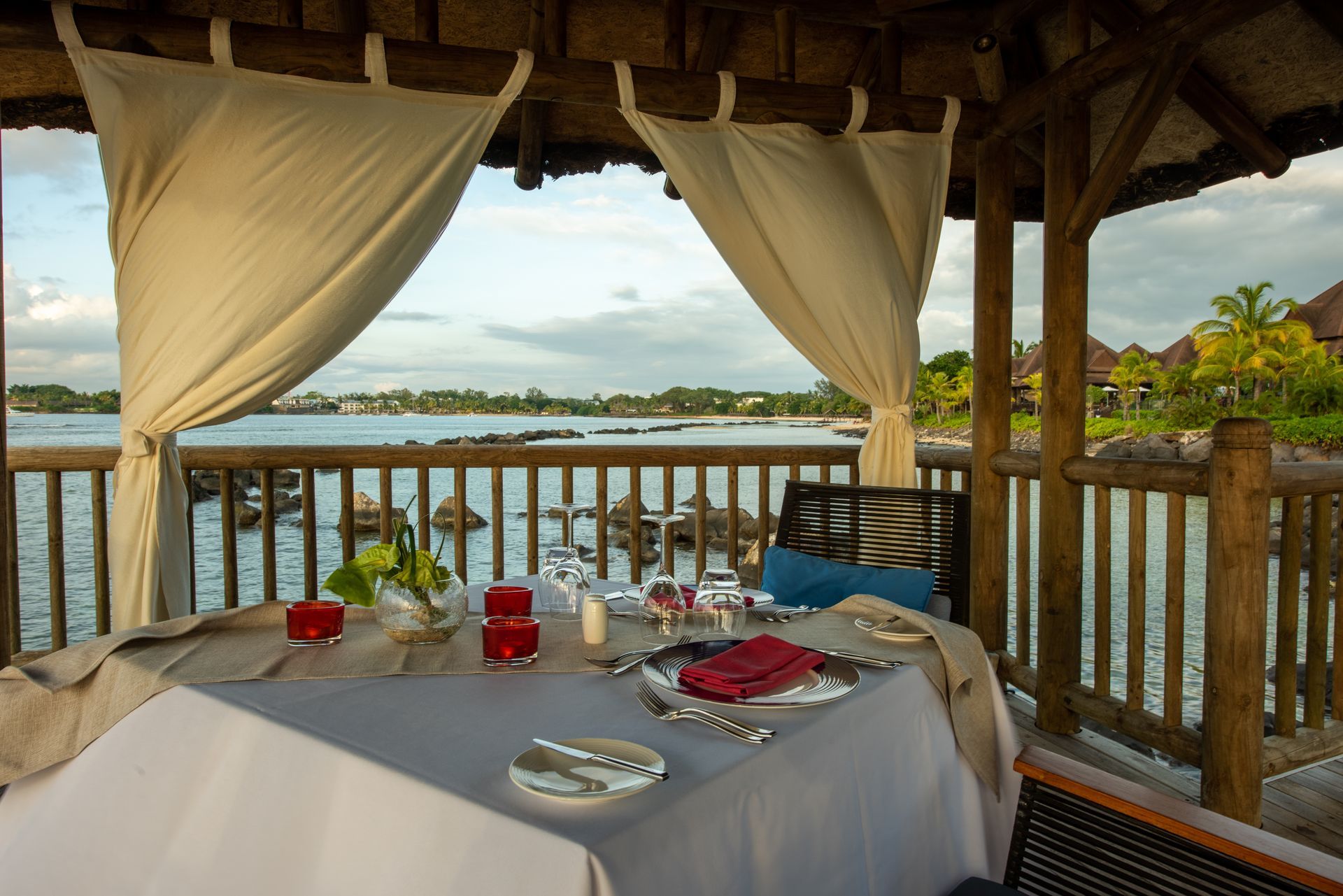 Table set for a meal overlooking water from a wooden gazebo, draped with white curtains.