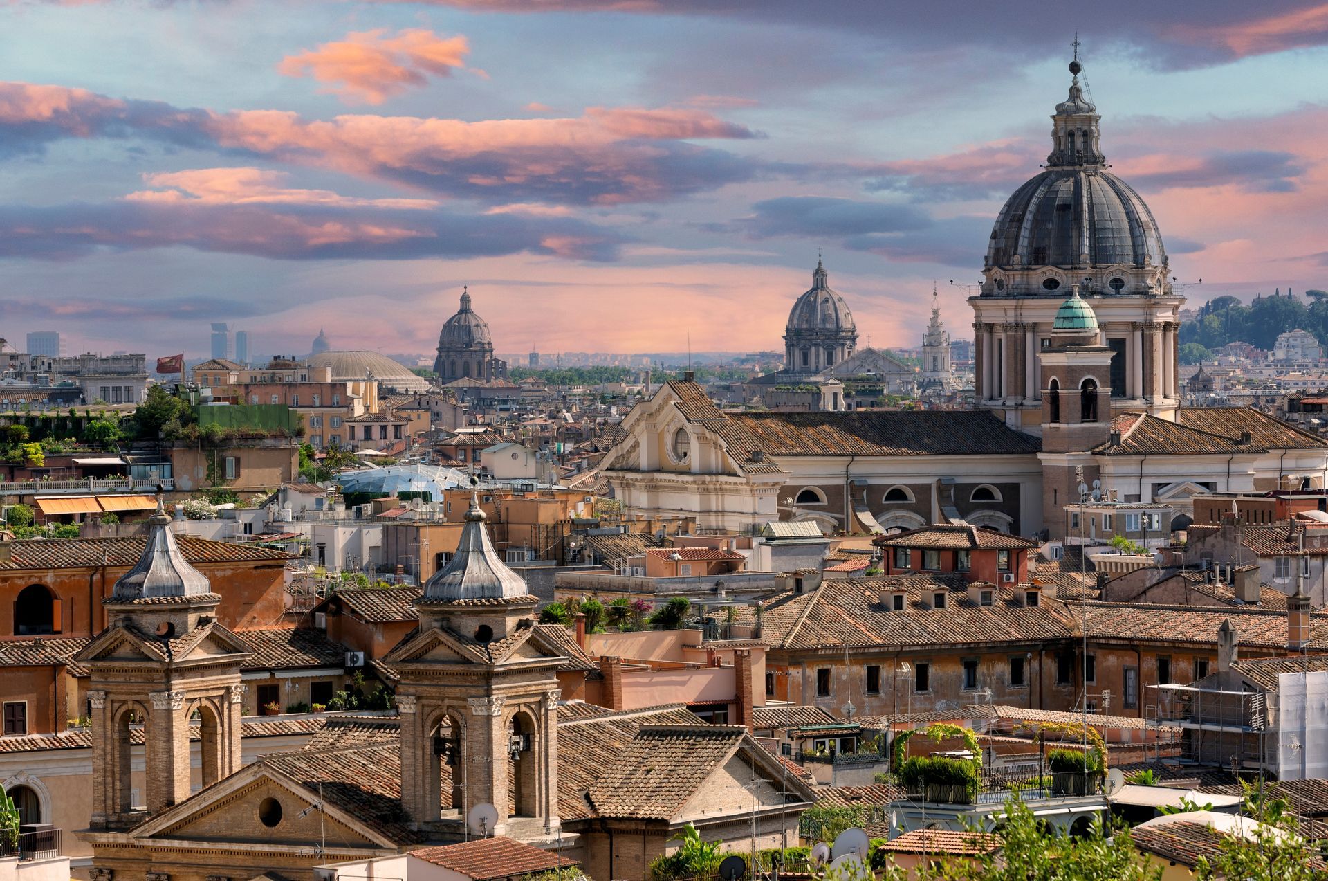 Skyline of Rome with several domed churches, rooftops, and a colorful sunset.