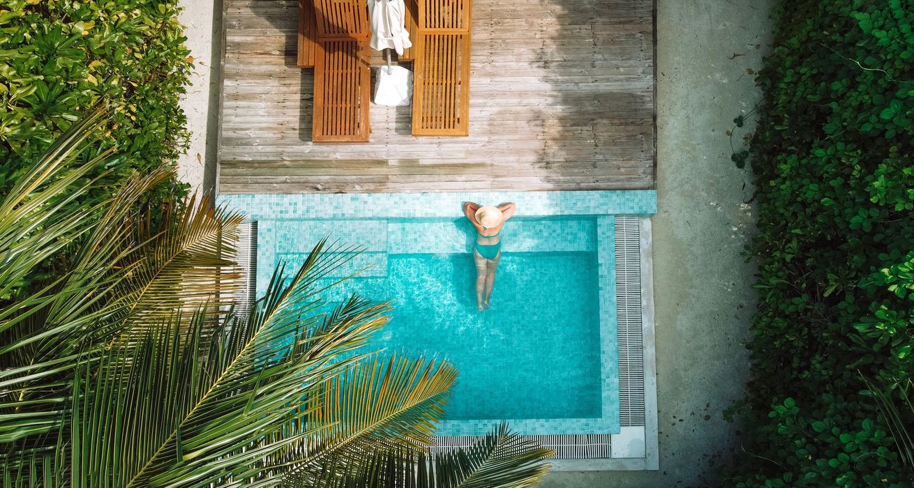 Man relaxing in a turquoise pool, aerial view. Wooden deck with chairs, palm trees.