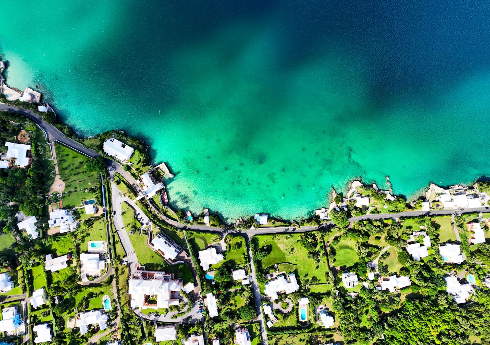 Aerial view of coastal houses with white roofs and green lawns next to turquoise and dark blue water.