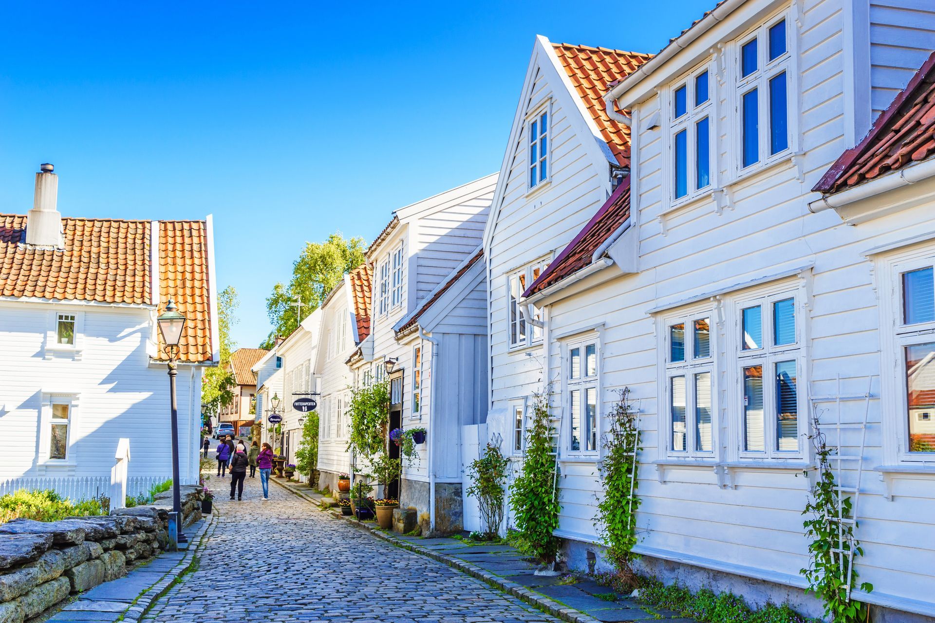 White wooden buildings line a cobblestone street under a clear blue sky; people walk along.