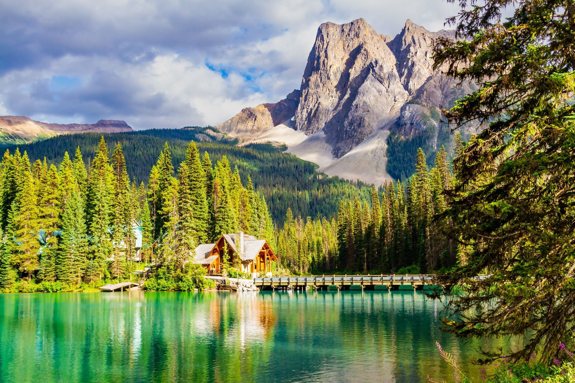 Emerald Lake with wooden bridge, lodge, lush green trees, and rocky mountains under a cloudy sky.