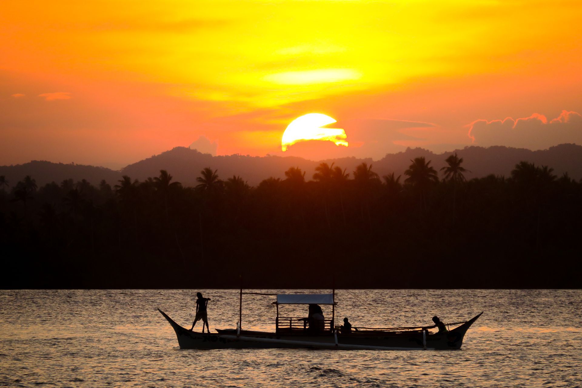 Boat silhouetted on water at sunset. Sun sets behind trees and mountains, coloring the sky orange.