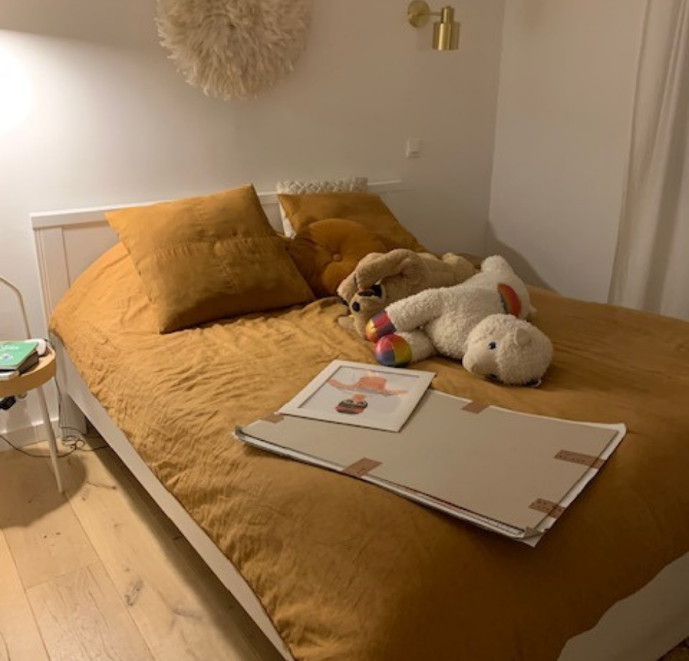 Lit avec draps orange, oreillers, peluches et œuvres d'art. Parquet blond et murs blancs.