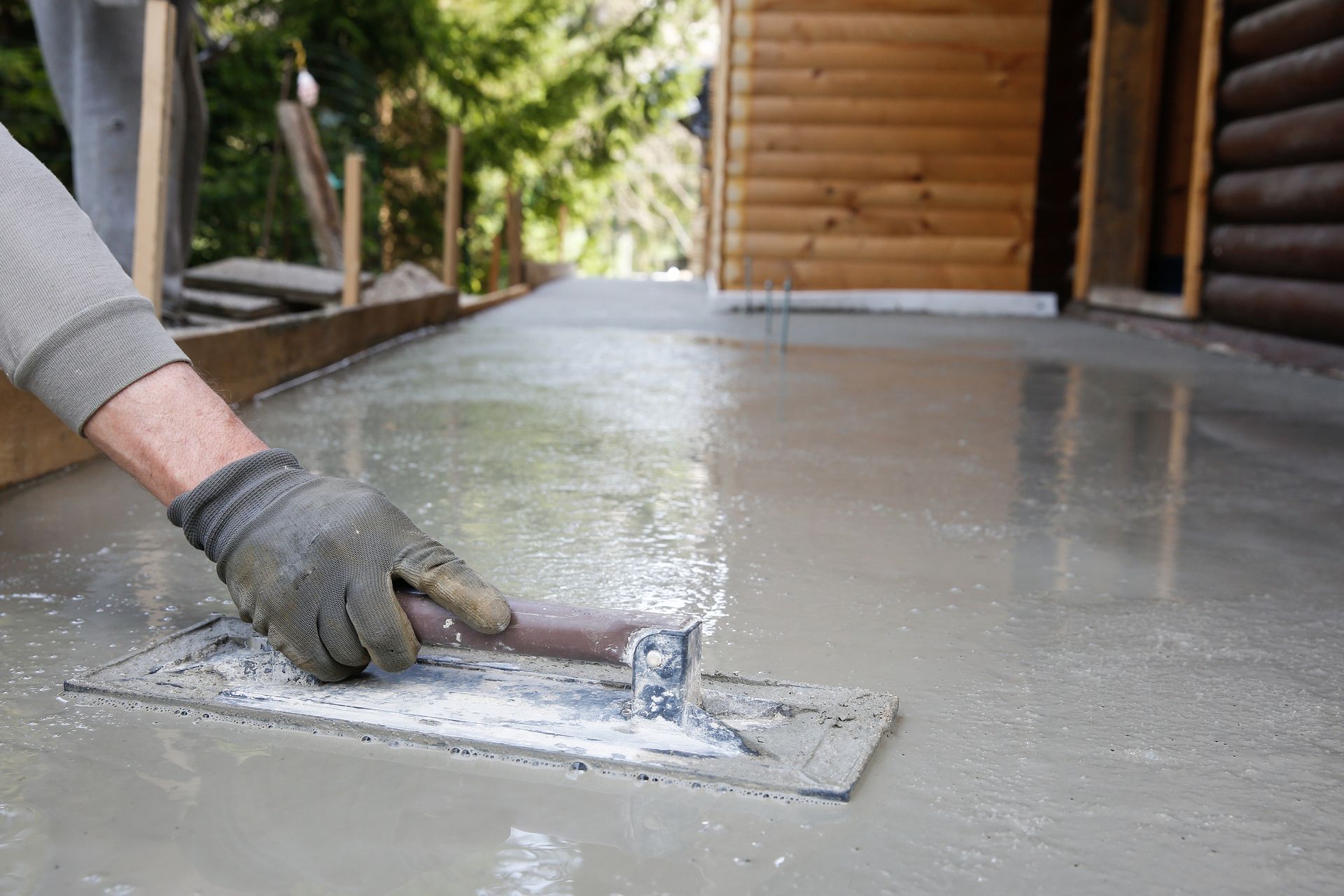 Main gantée lissant un sol en béton humide à la truelle, à côté d'un bâtiment en bois
