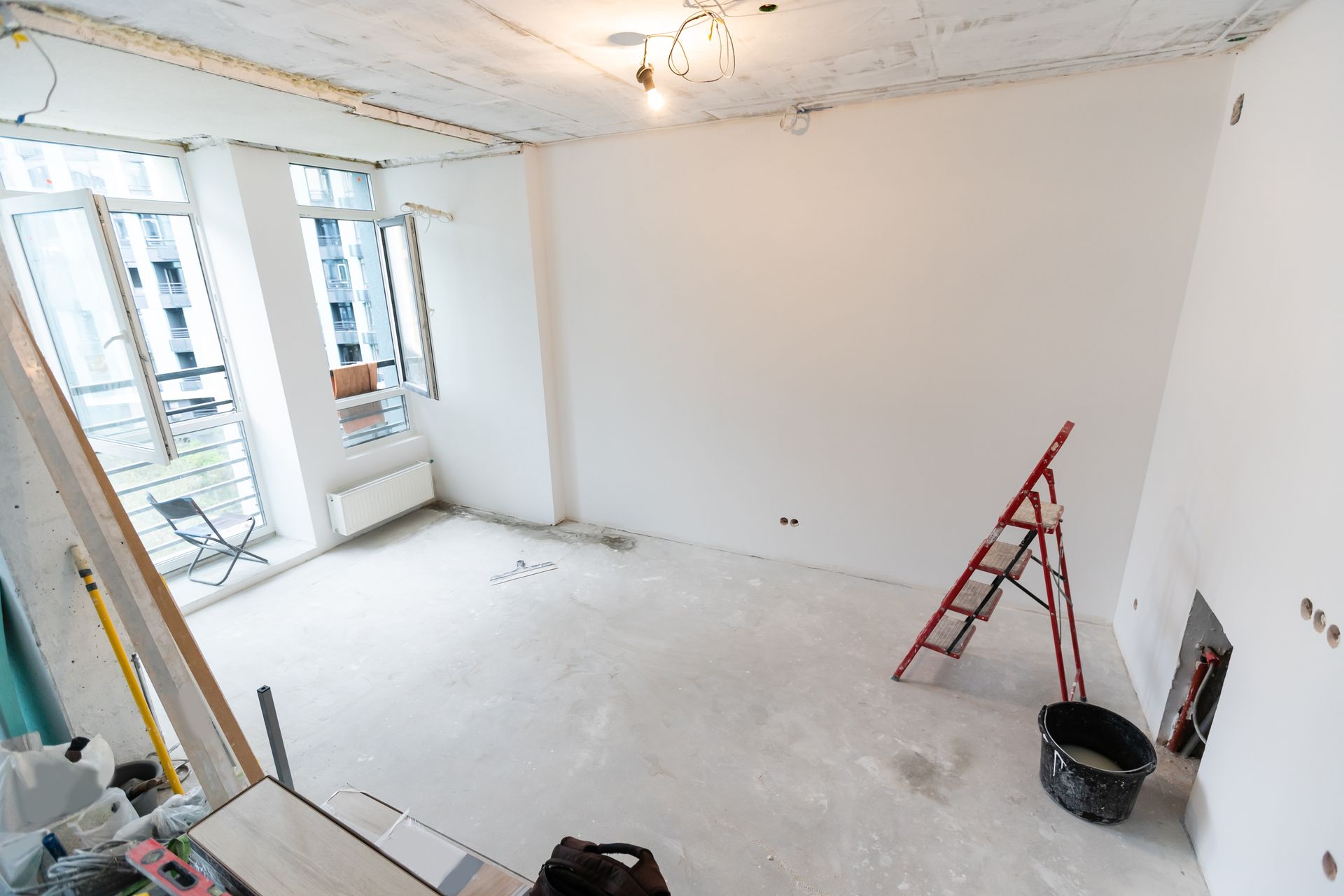 Pièce blanche vide en cours de rénovation, avec une échelle rouge, un seau et des fenêtres le long d'un mur.