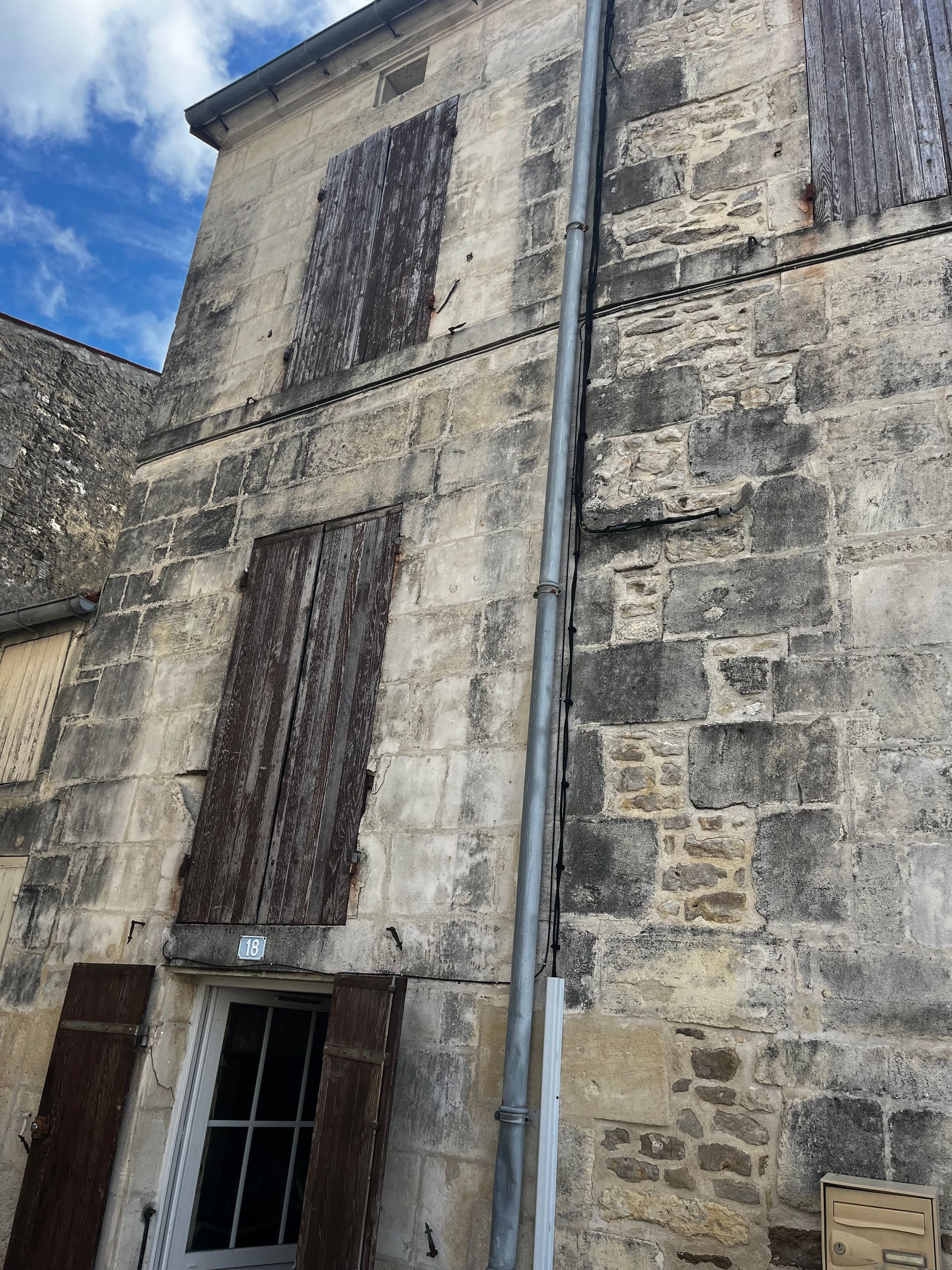 Façade d'un bâtiment en pierre, avec fenêtres à barreaux et descente de gouttière métallique, se détachant sur un ciel bleu.