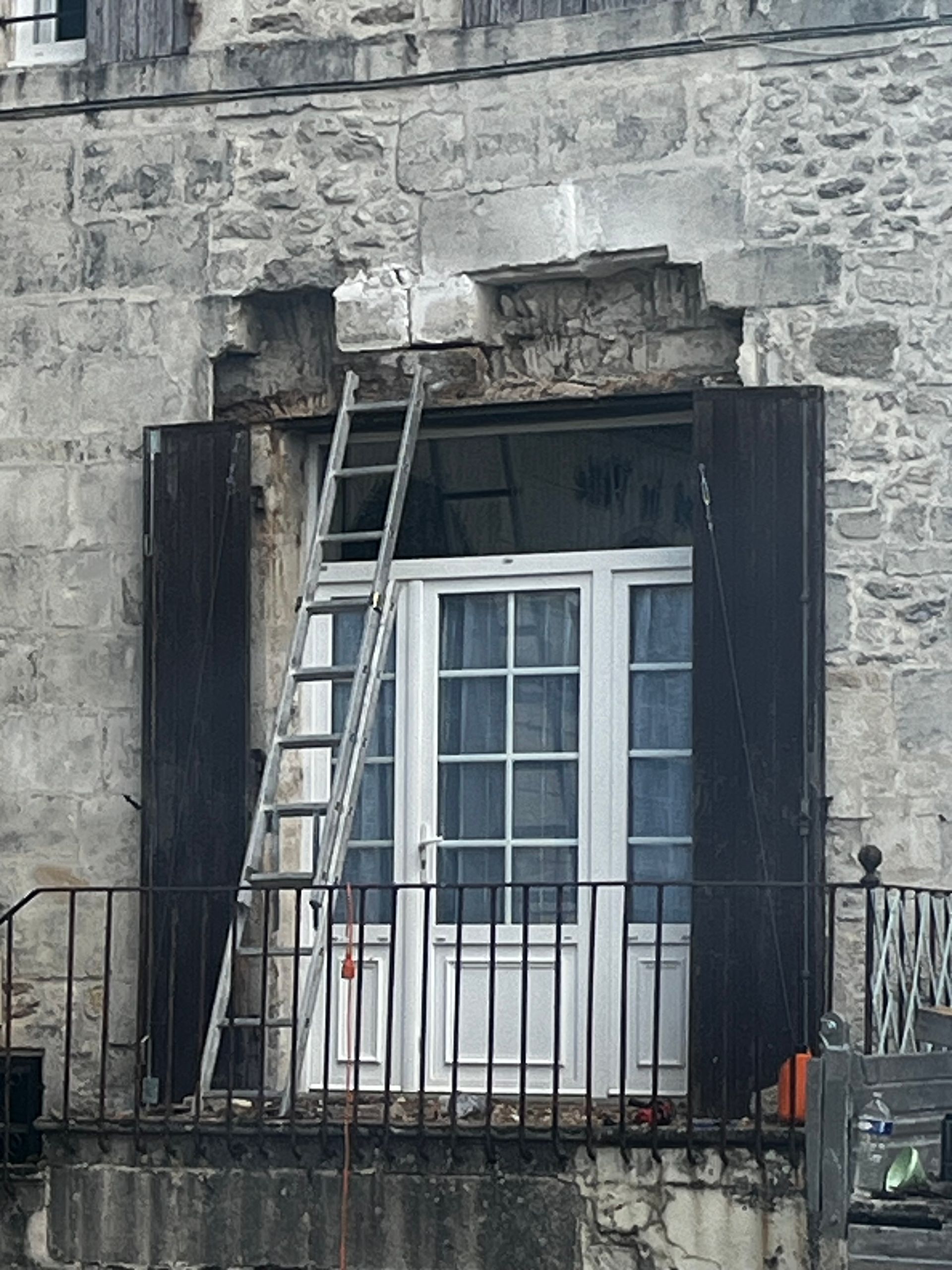 Une échelle est appuyée contre un mur de pierre au-dessus d'une porte blanche à volets noirs et à rambarde métallique.