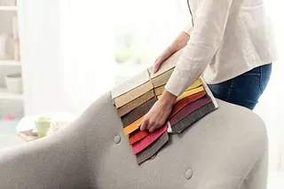 Mujer sosteniendo muestras de tela, comparando colores con un diván gris en la sala de estar.