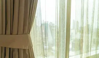 Cortinas beige, una de ellas recogida con un lazo, que deja ver una vista de la ciudad a través de una cortina transparente.