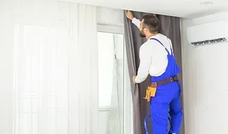 Hombre con mono azul en una escalera colgando una cortina gris en una habitación blanca con una ventana.