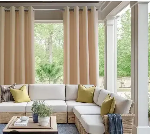 Cortinas de exterior de color crema enmarcan un sofá seccional con almohadas amarillas en un patio.