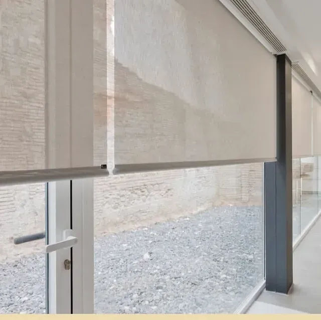 Ventana con persianas enrollables de color beige, parcialmente bajadas, con vista a un espacio exterior.