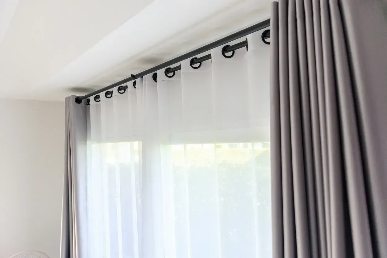 Cortinas grises y blancas transparentes colgadas de una barra negra sobre una ventana en una habitación luminosa.