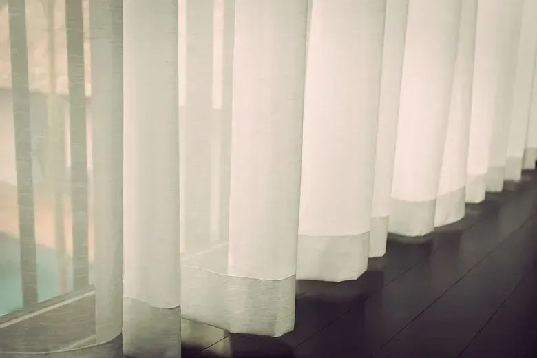 Cortinas blancas transparentes colgando frente a una ventana, proyectando sombras suaves sobre el piso de madera oscura.