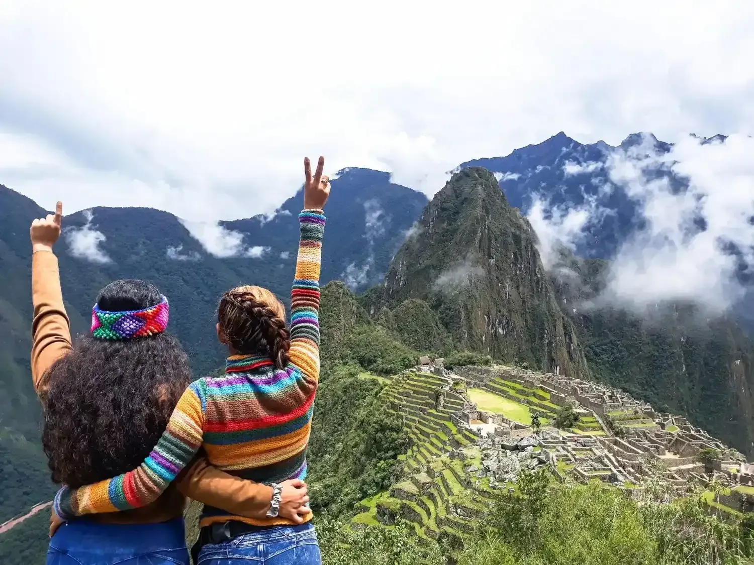 Dos personas con los brazos levantados, vista Machu Picchu, montañas, cielo nublado.