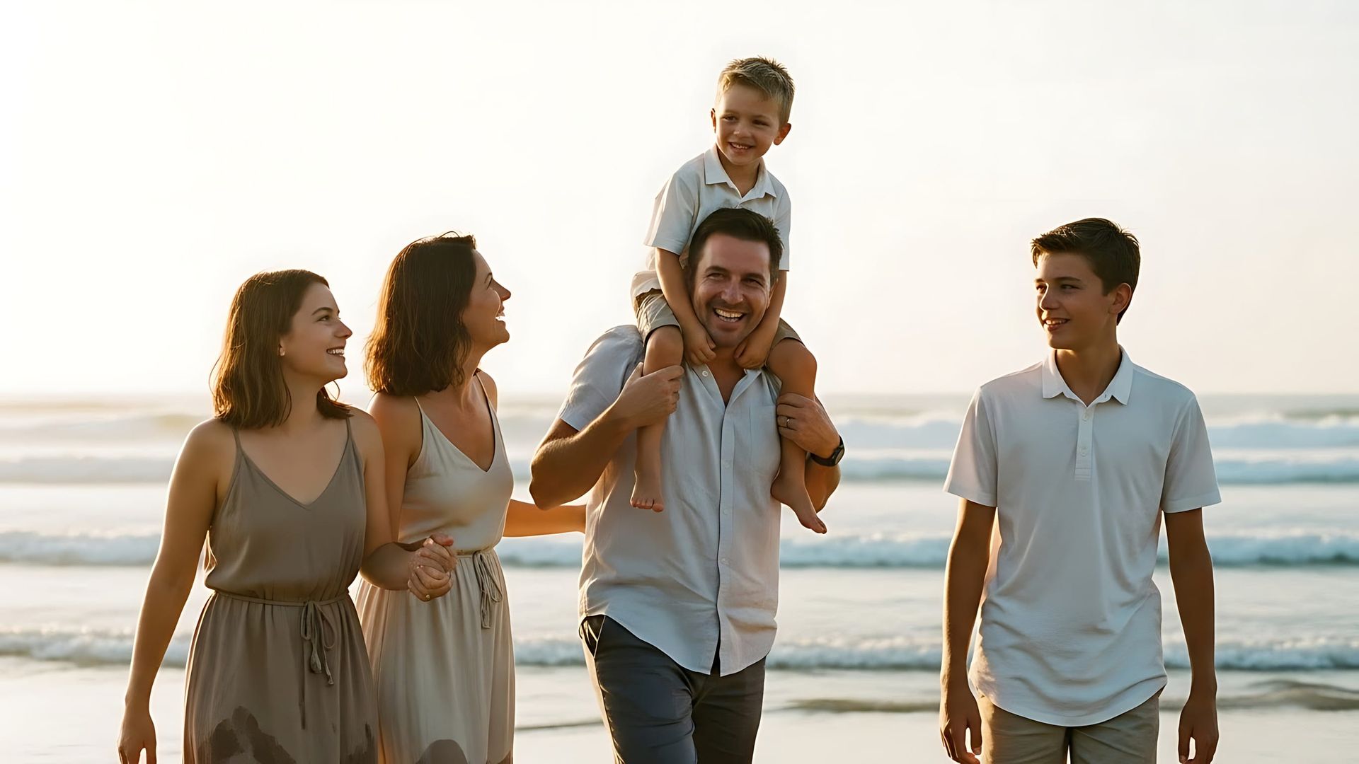Familia caminando por una playa; padre cargando al niño en hombros, sonriendo; día soleado.