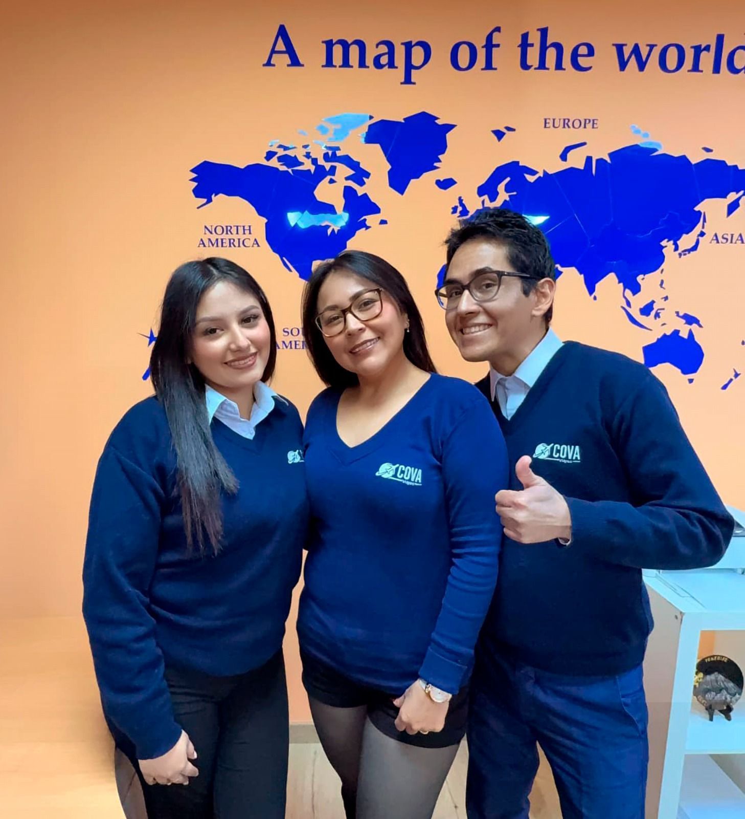 Tres personas con suéteres azules posan frente a un mapa del mundo. Una levanta el pulgar.