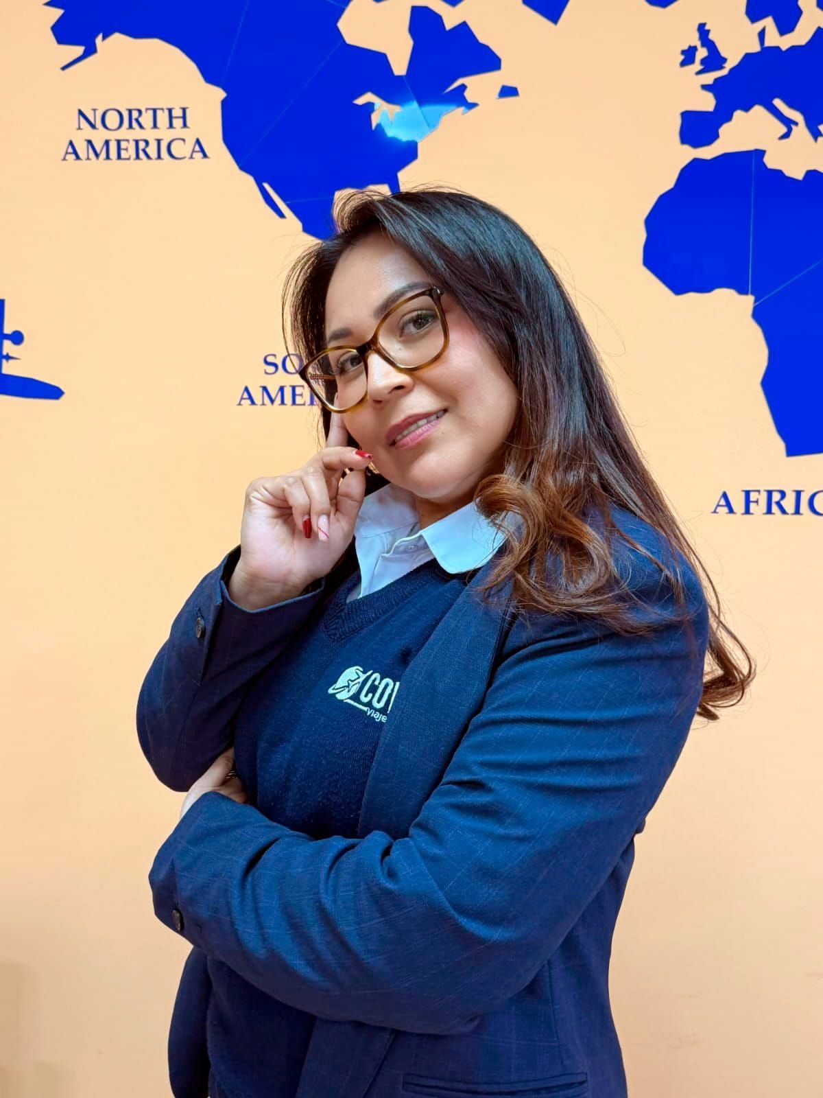 Mujer con blazer azul, gafas, posando delante de un mapa del mundo, con el dedo en la barbilla.