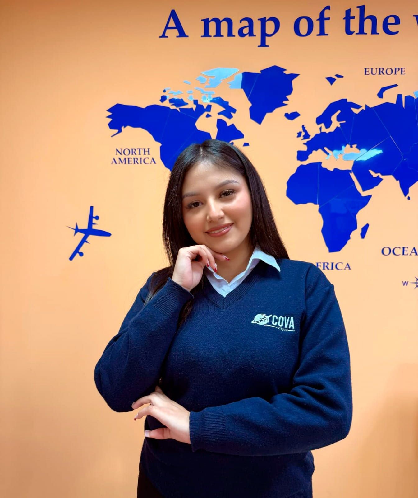 Una mujer con un suéter azul está cerca de un mapa del mundo, sonriendo y mirando a la cámara.