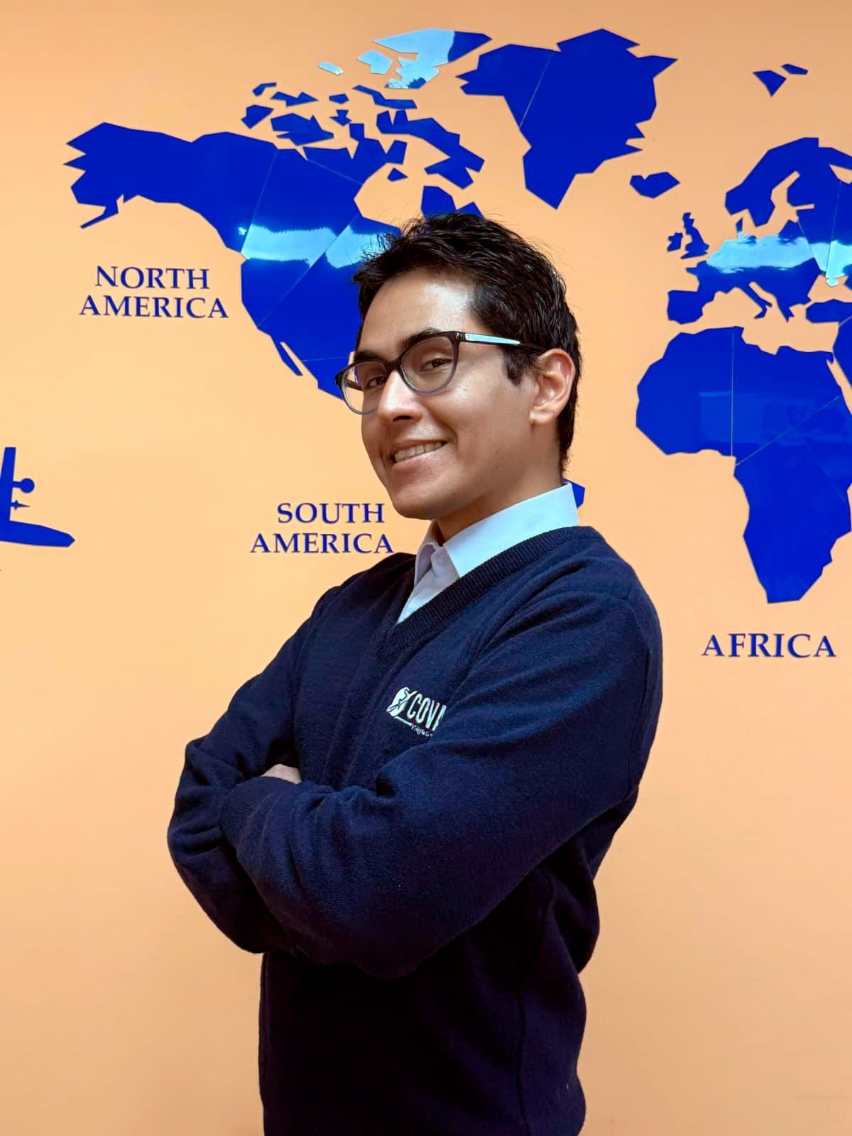 Hombre con gafas, con los brazos cruzados, de pie frente a un mapa del mundo en una pared naranja.