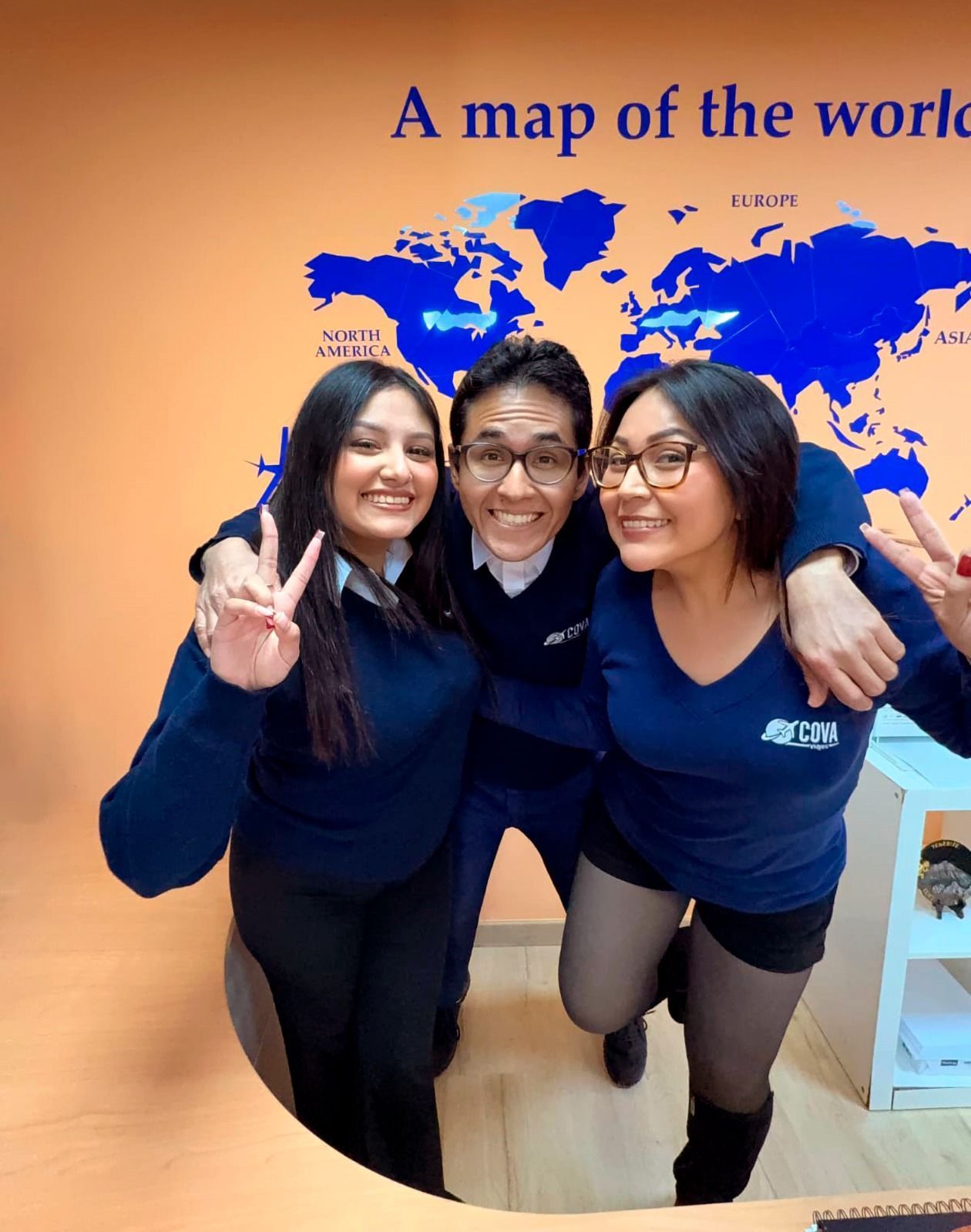 Tres personas sonrientes con camisas azules posan frente a un mapa del mundo, haciendo señales de paz.