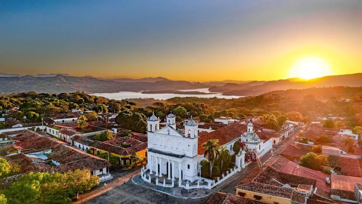 Puesta de sol sobre una iglesia blanca con techos de tejas rojas, con vistas a una ciudad y un lago.
