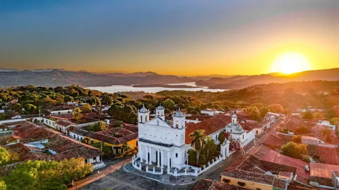 Puesta de sol sobre una iglesia blanca con techos de tejas rojas, con vistas a una ciudad y un lago.