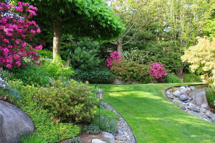 Un jardin luxuriant avec une pelouse d'un vert éclatant, des arbustes à fleurs rose vif, de gros rochers d'aménagement paysager et des arbres matures.